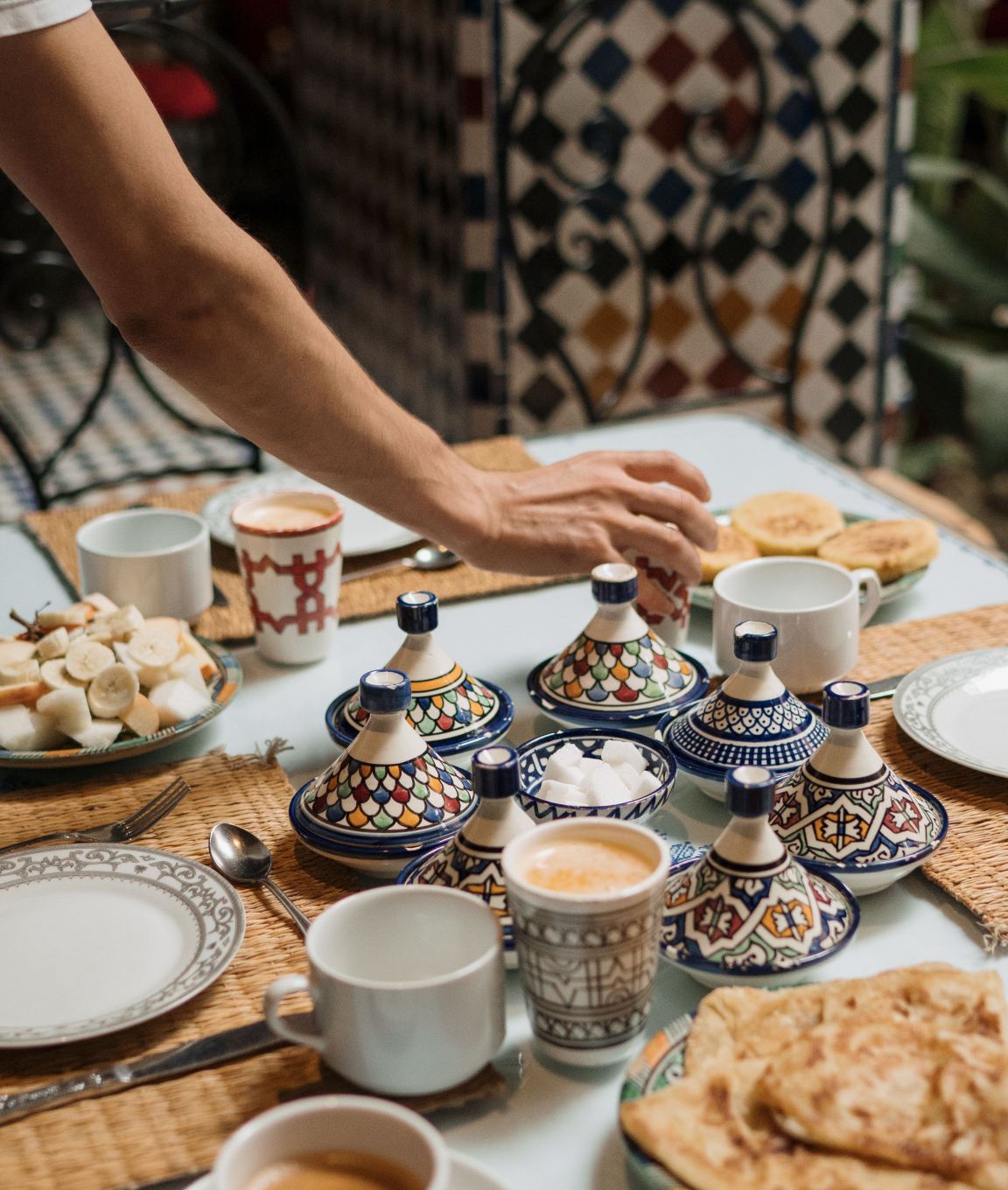 A hand reaching across a breakfast table set with blue-and-white teapots and bread
