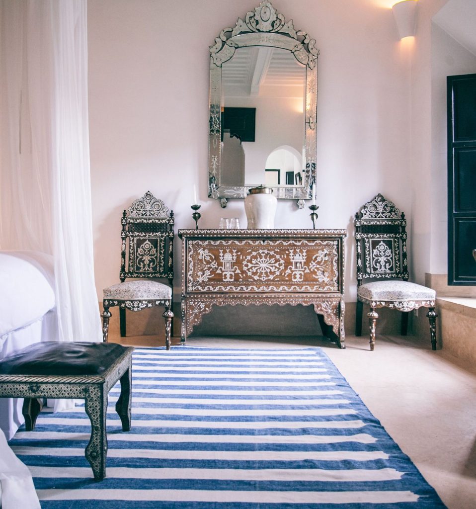 A white bedroom with a mirrored console, inlaid chairs and a blue-striped rug