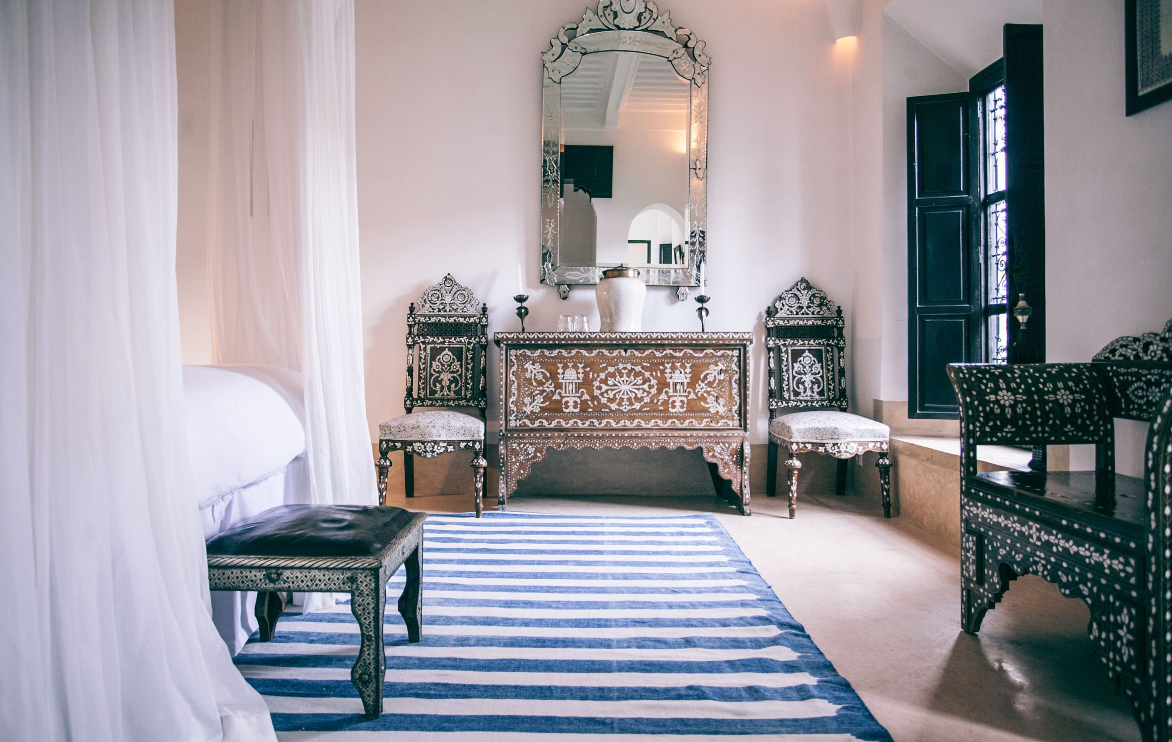 A white bedroom with a mirrored console, inlaid chairs and a blue-striped rug