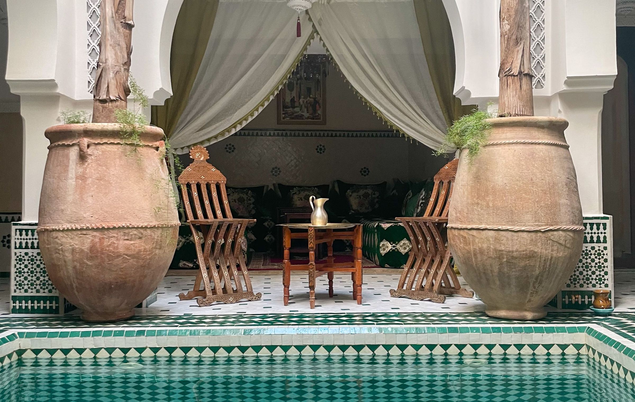 Two tall clay jars and wooden chairs beside a tiled pool