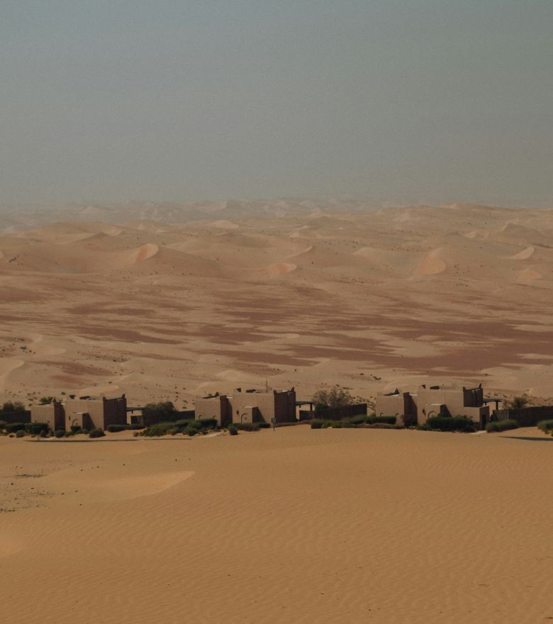 A row of earth-built houses below wide sand dunes under a hazy sky