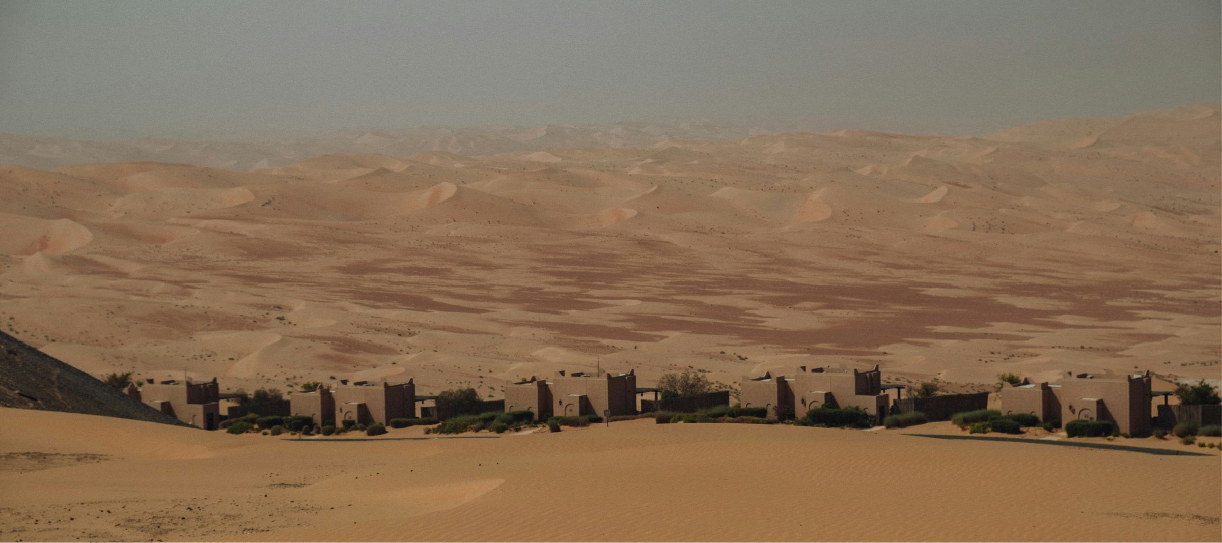 A row of earth-built houses below wide sand dunes under a hazy sky