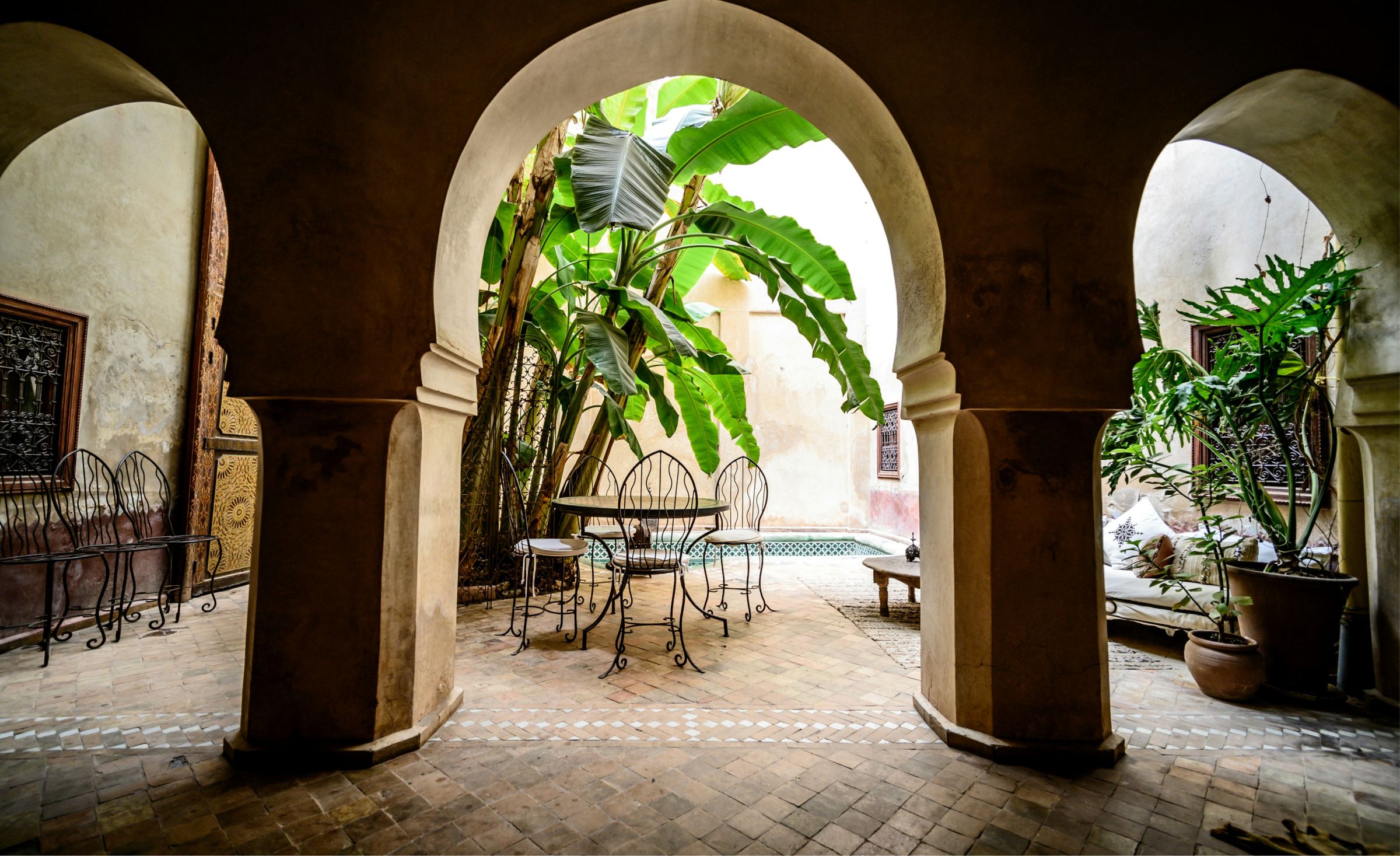 A table and iron chairs under a horseshoe arch, banana leaves behind