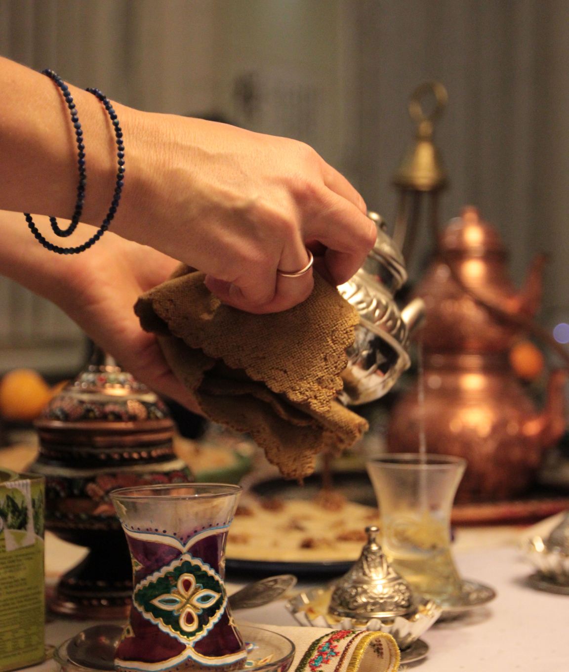 Hands lifting a brass teapot over a table with tea glasses