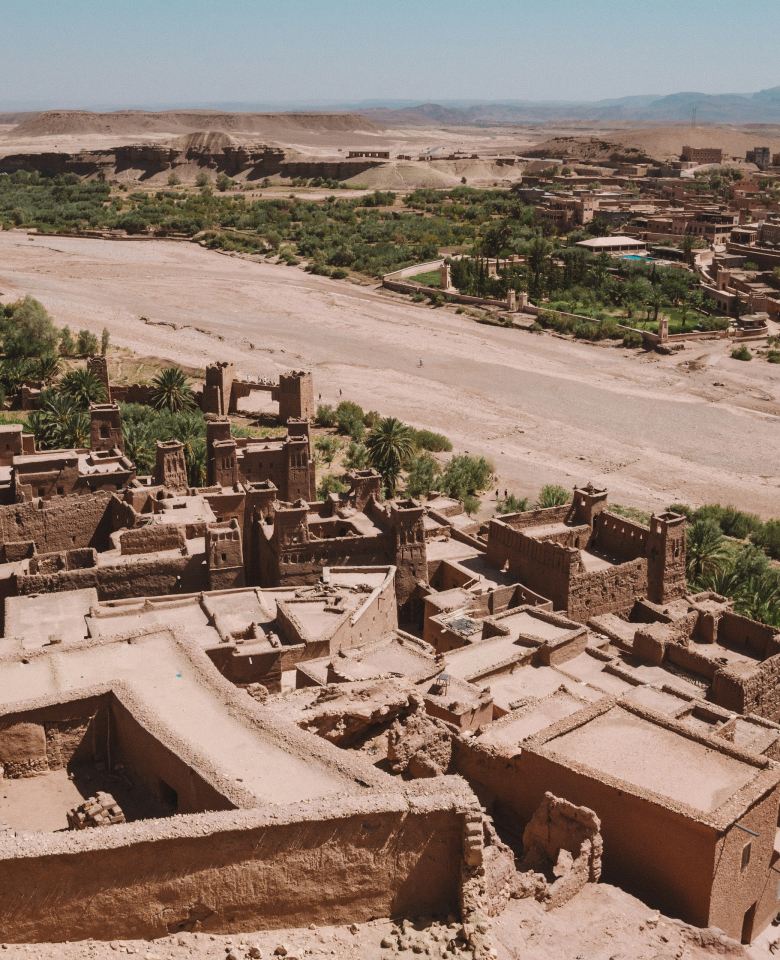 Earth rooftops of Aït Ben Haddou above a dry riverbed, palm groves and desert hills