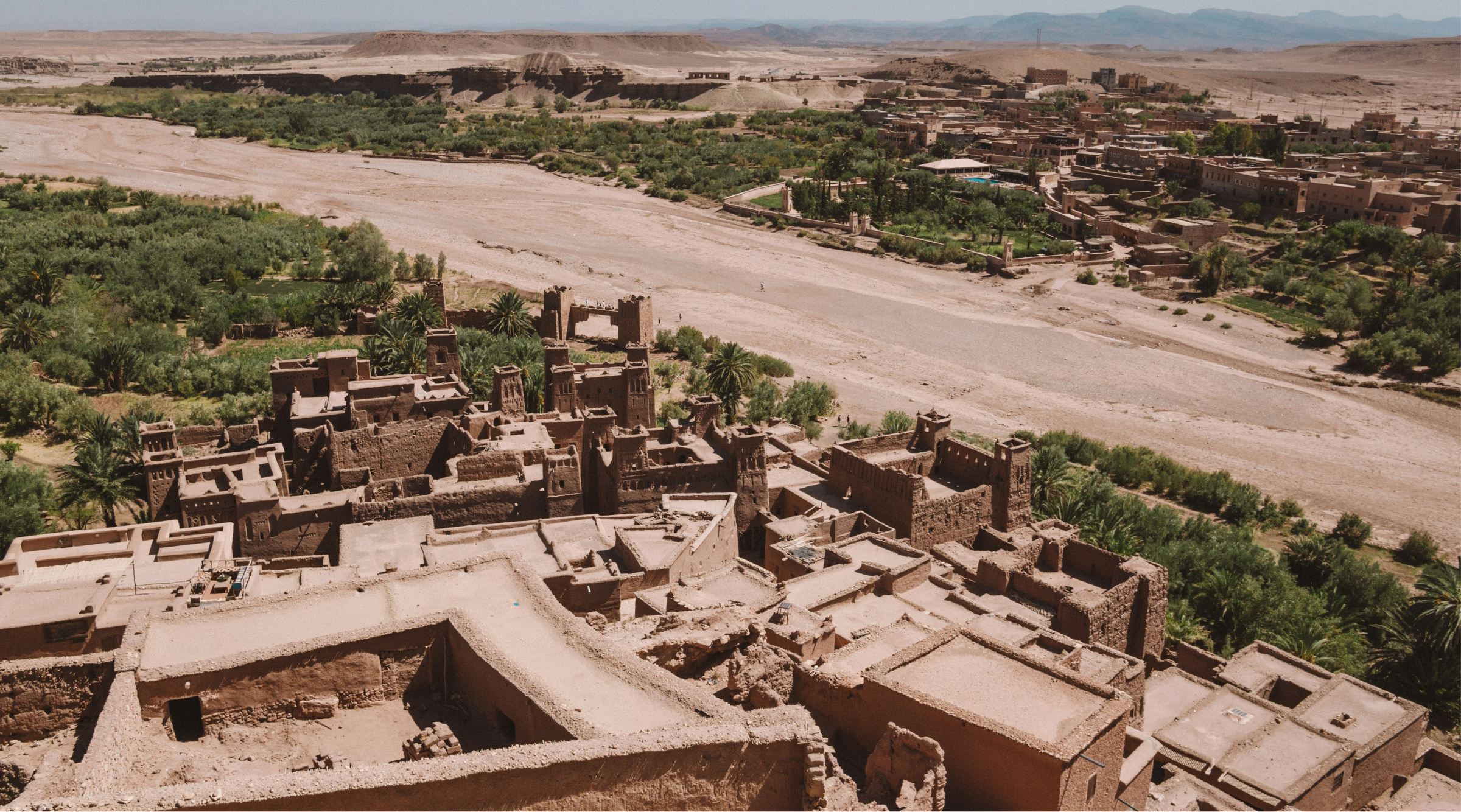 Earth rooftops of Aït Ben Haddou above a dry riverbed, palm groves and desert hills